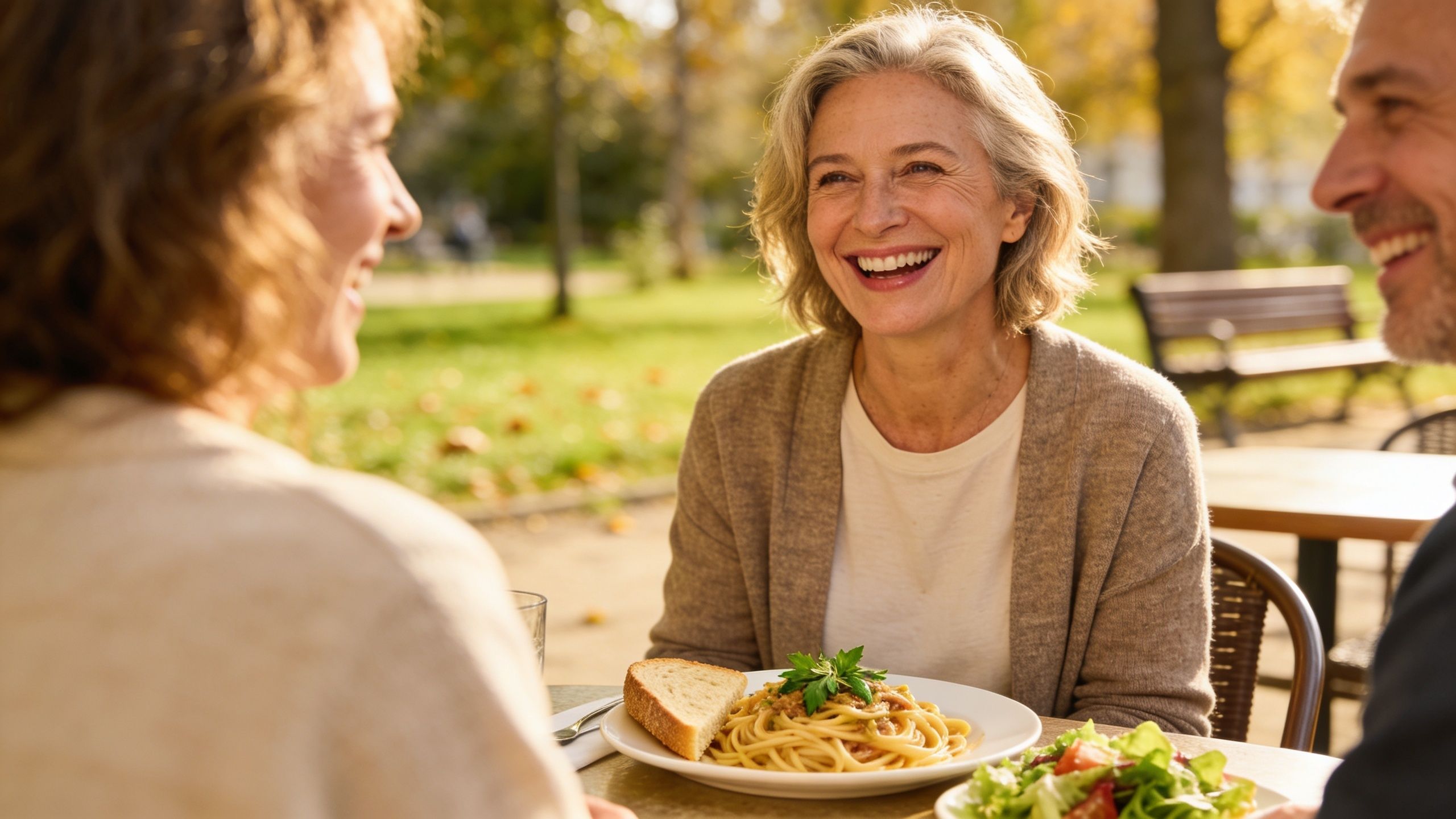 A middle-aged woman laughing while enjoying a healthy lunch with her friends in a sunny outdoor setting.
